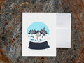 A greeting card with a snow globe illustration with a cabin, skier, and two dogs among a snowy landscape, displayed on a wooden surface with an envelope.