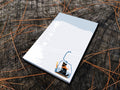 Notepad with illustration of a dog with goggles and orange scarf riding a ski lift, placed on a wooden surface.