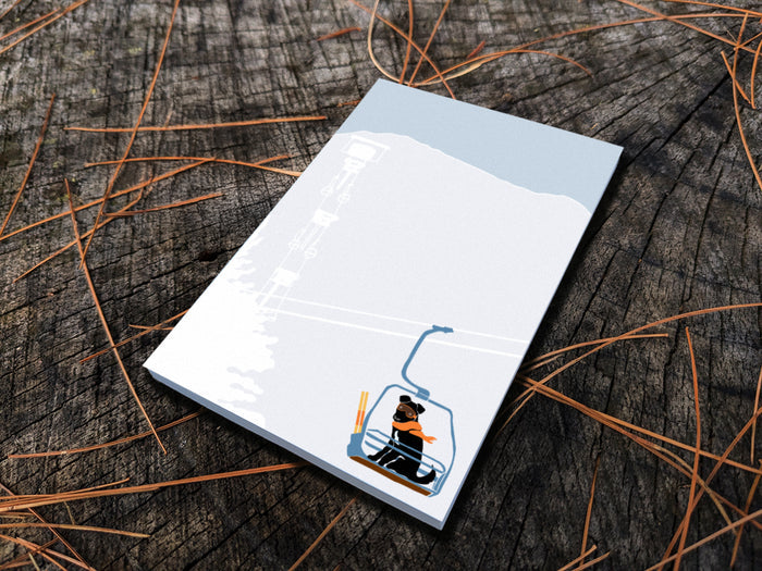 Notepad with illustration of a dog with goggles and orange scarf riding a ski lift, placed on a wooden surface.
