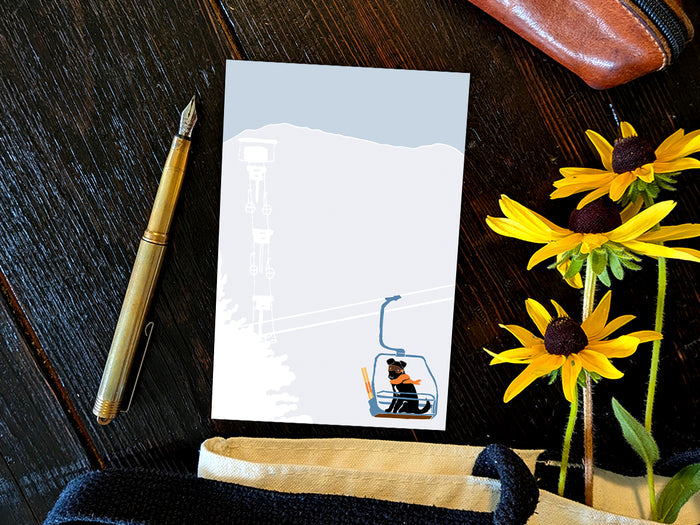 Notepad with illustration of a dog with goggles and orange scarf riding a ski lift, placed on a wooden surface with a gold pen, and yellow flowers.