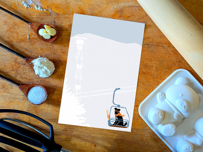 Notepad with illustration of a dog with goggles and orange scarf riding a ski lift, placed on a wooden surface with baking supplies.