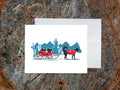 Greeting card with dog in a red jacket pulling a sled with another dog in a wintry landscape, on a wooden surface with white envelope.