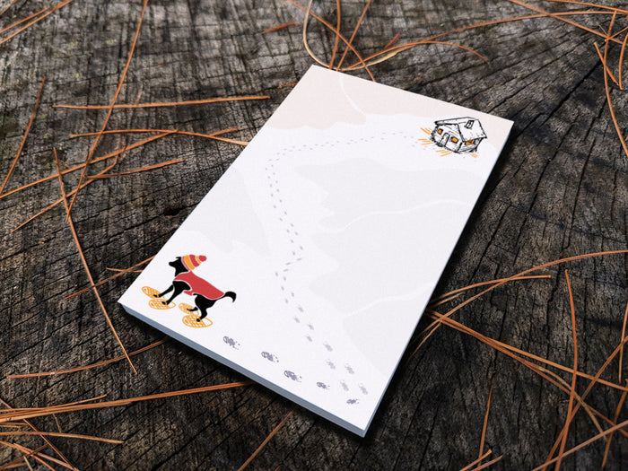 Notepad with illustration of a black dog with winter hat, red jacket, and snowshoes, with a trail a footprints leading from a rustic cabin in the background, placed on a wooden surface.
