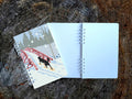 A spiral-bound notebook with an illustration of a dog snowshoeing over a bridge, shown on top of an open blank version of the notebook on a wood surface.
