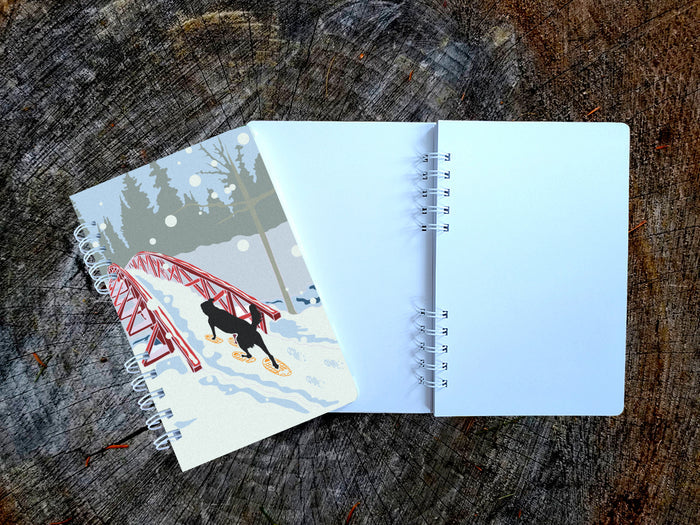 A spiral-bound notebook with an illustration of a dog snowshoeing over a bridge, shown on top of an open blank version of the notebook on a wood surface.