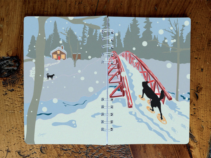 A spiral-bound notebook with an illustration of a dog snowshoeing over a bridge toward a cabin and another dog, shown on a wood surface.