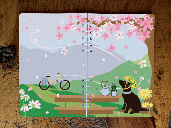 Spiral-bound notebook with a scenic illustration of a dog, bicycle, and picnic table on a wooden surface.