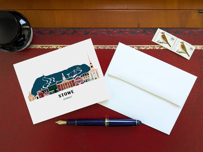 Greeting card with image of buildings arranged in a city skyline with the text Stowe Vermont on a table with blue pen and envelope and stamps.