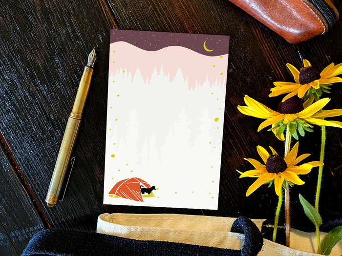 Notepad with an illustration of a black dog in red tent camping under night sky with fireflies, placed on a wood surface with a gold pen, and yellow flowers.