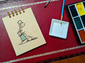 Notebook with a drawing of coffee brewing on a rock, next to a paint palette and brush on a red surface.