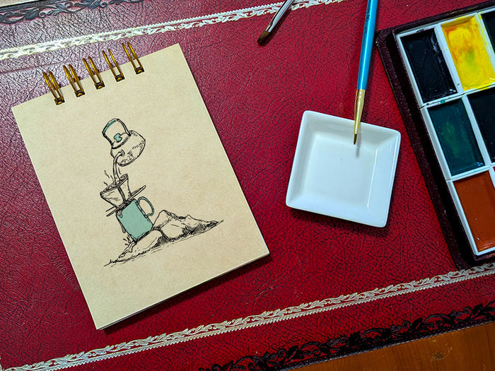 Notebook with a drawing of coffee brewing on a rock, next to a paint palette and brush on a red surface.