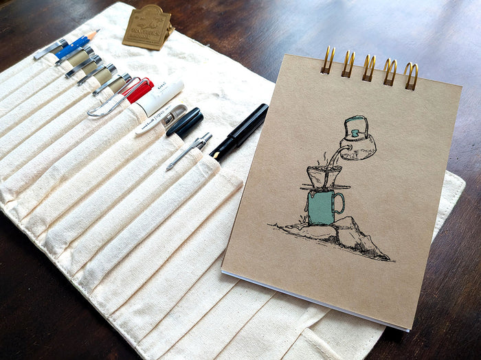 Notebook with a drawing of coffee brewing on a rock, next a pen case on a wood surface.
