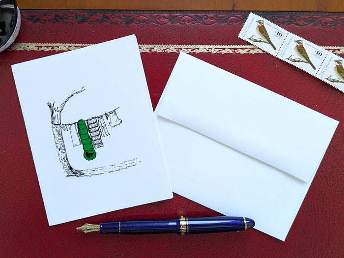 White card with a black ink drawing of laundry hanging from a tree on a red surface with a pen, envelope and stamp.