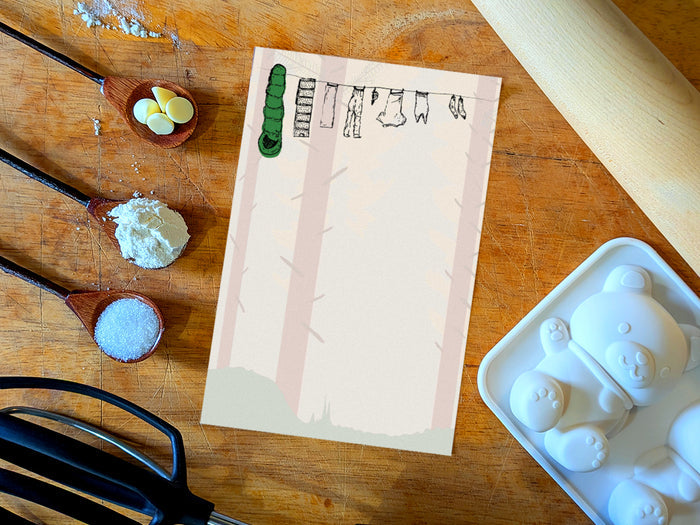 Notepad with illustration of clothes and sleeping bag hanging on a line in between pine trees in a forest, placed on wooden surface with baking supplies.
