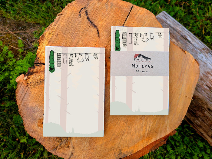Two notepads with illustration of clothes and sleeping bag hanging on a line in between pine trees in a forest, placed on wooden surface.