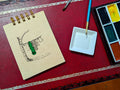 Notebook with a drawing of clothes and sleeping bag hanging on line, next to a paint palette and brush on a red surface.