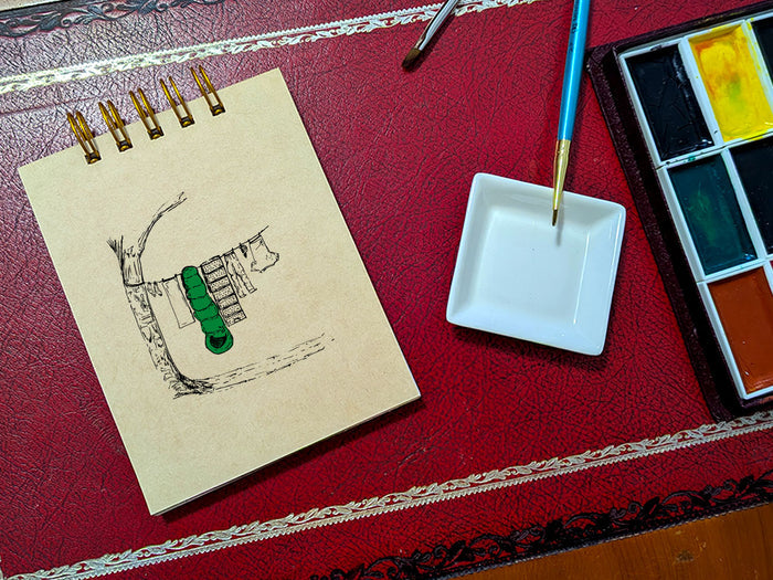 Notebook with a drawing of clothes and sleeping bag hanging on line, next to a paint palette and brush on a red surface.