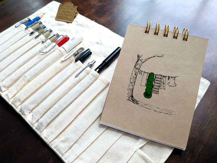 Notebook with a drawing of clothes and sleeping bag hanging on line, next a pen case on a wood surface.