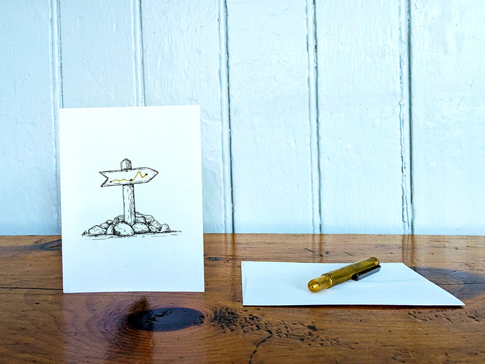 White card with a black ink drawing of a trail marker on a wooden surface with a pen and envelope.