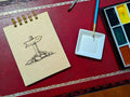 Notebook with a drawing of a trail marker, next to a paint palette and brush on a red surface.
