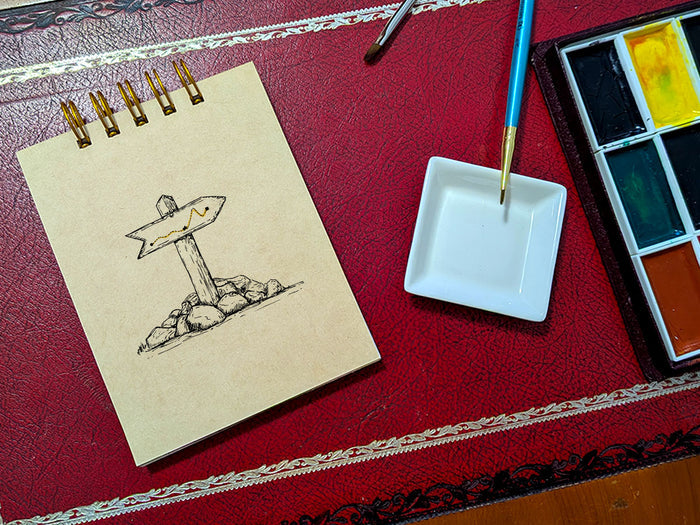 Notebook with a drawing of a trail marker, next to a paint palette and brush on a red surface.
