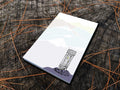 Notepad with an illustration of an outhouse in the mountains, placed on a wood surface.