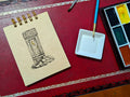 Notebook with a drawing of an outhouse, next to a paint palette and brush on a red surface.