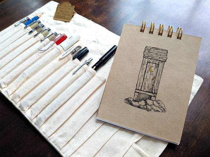 Notebook with a drawing of an outhouse, next a pen case on a wood surface.