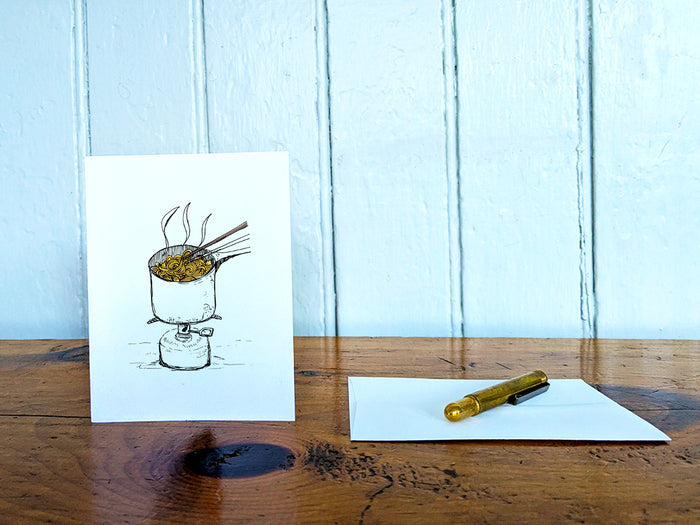 White card with a black ink drawing of a pot of ramen over a camp stove on a wooden surface with a pen and envelope.
