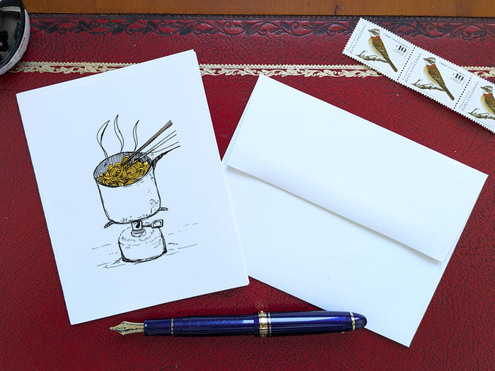 White card with a black ink drawing of a pot of ramen over a camp stove on a red surface with a pen, envelope and stamp.