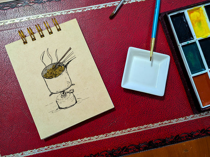 Notebook with a drawing of ramen cooking on a camp stove, next to a paint palette and brush on a red surface.