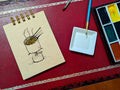 Notebook with a drawing of ramen cooking on a camp stove, next to a paint palette and brush on a red surface.