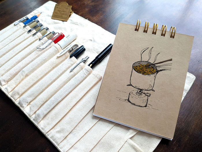 Notebook with a drawing of ramen cooking on a camp stove, , next a pen case on a wood surface.