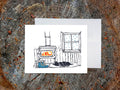 A greeting card with a illustrated scene featuring a wood-burning stove, snow covered window, sleeping dog, and hiking boots, displayed on a wooden background with an envelope behind it.