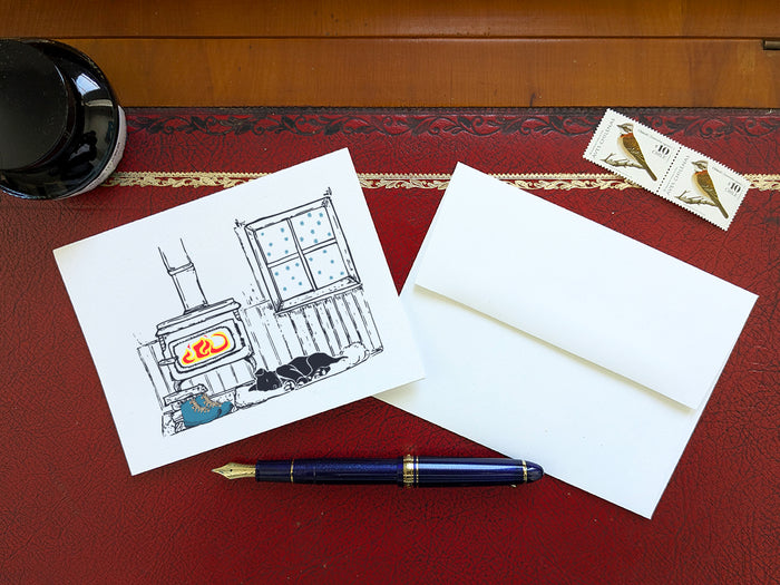A greeting card with a illustrated scene featuring a wood-burning stove, snow covered window, sleeping dog, and hiking boots, displayed on a red desk with an envelope, blue fountain pen and stamps.